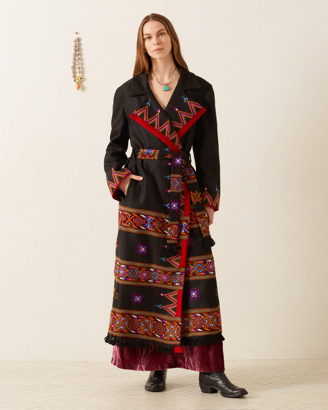 A woman stands against a plain wall, wearing the Penelope Wildflower Wool Coat, featuring vibrant Western-inspired geometric embroidery and a matching belt. Her coat complements her long hair and ankle boots. With a calm expression, she stands by a decorative hanging on the wall beside her.