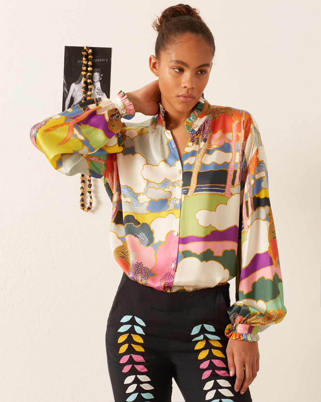 A person wearing a Poet Aurora Silk Blouse with colorful patterns and black pants stands in a room with a neutral background.