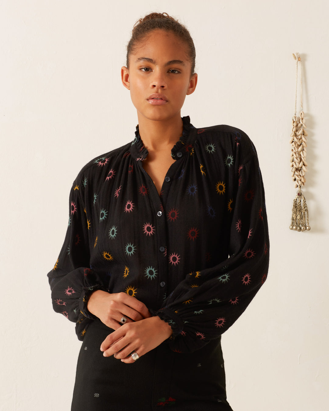 A person stands against a plain wall, wearing the Poet Evening Star Blouse adorned with a cosmic print and black pants. A small decorative item hangs on the wall, enhancing the celestial vibe.