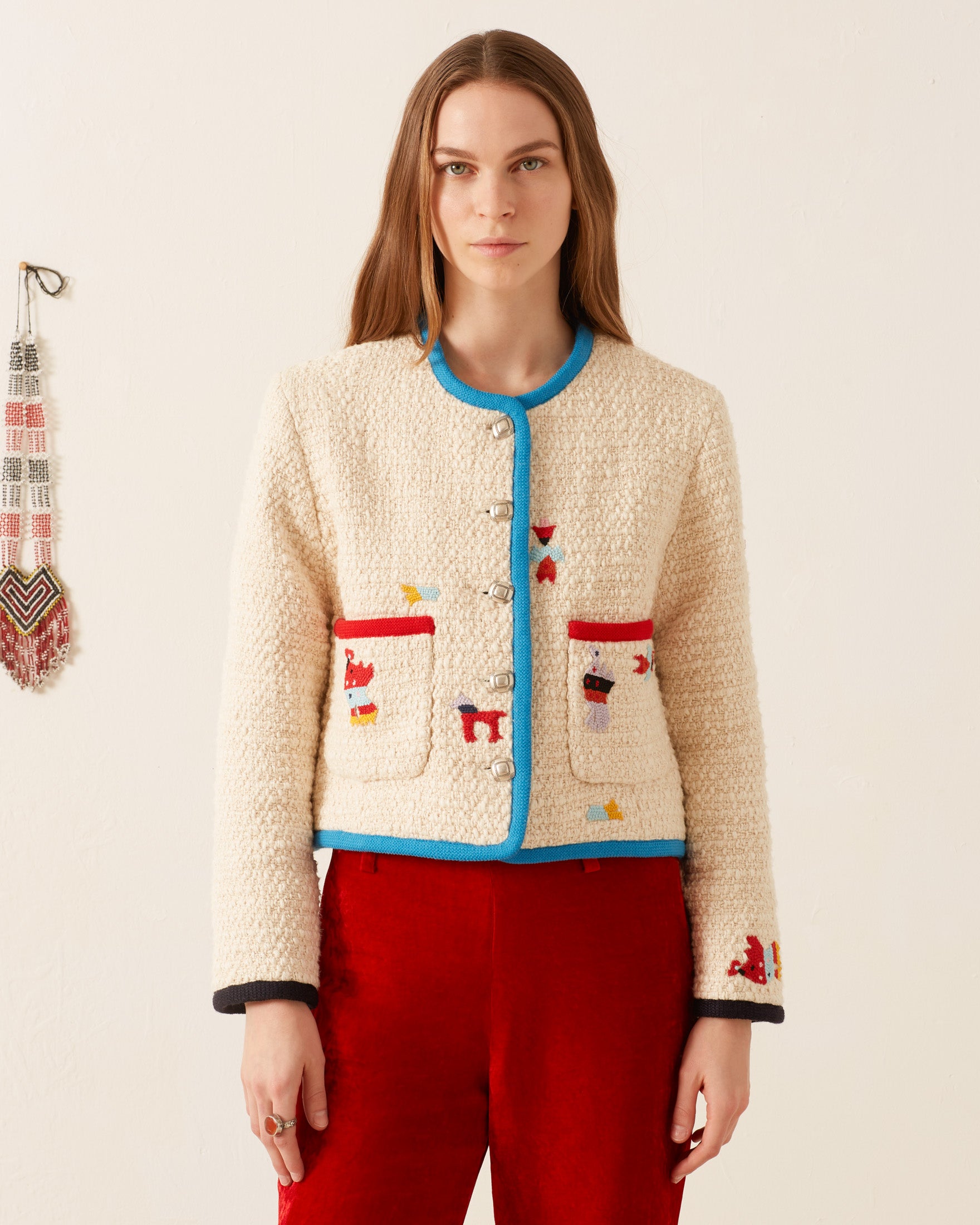 A person wearing the Rowan Pearl Boucle Jacket, which is cream with red and blue accents and lined with cotton-silk, stands against a plain background. The jacket features several colorful embroidered designs. They are also wearing red pants. A beaded decoration hangs on the wall to the left.