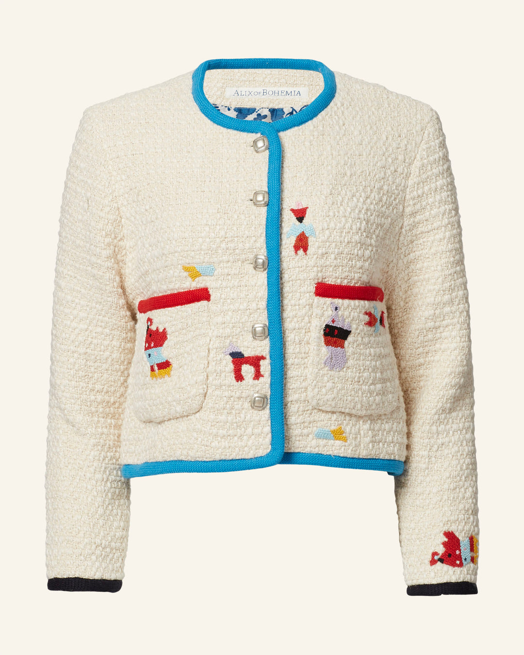 The Rowan Pearl Boucle Jacket is a white bouclé jacket with blue trim, adorned with colorful and whimsical embroidery, including red pockets and abstract shapes. It features pearl-like buttons and long sleeves.