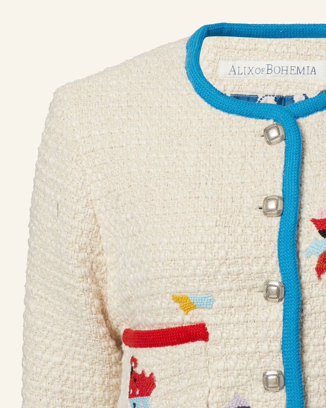 Close-up of the Rowan Pearl Boucle Jacket, featuring vibrant embroidered motifs with geometric shapes in red, blue, and yellow. Enhanced with blue trim and silver buttons, the top left corner reads "Alix of Bohemia.