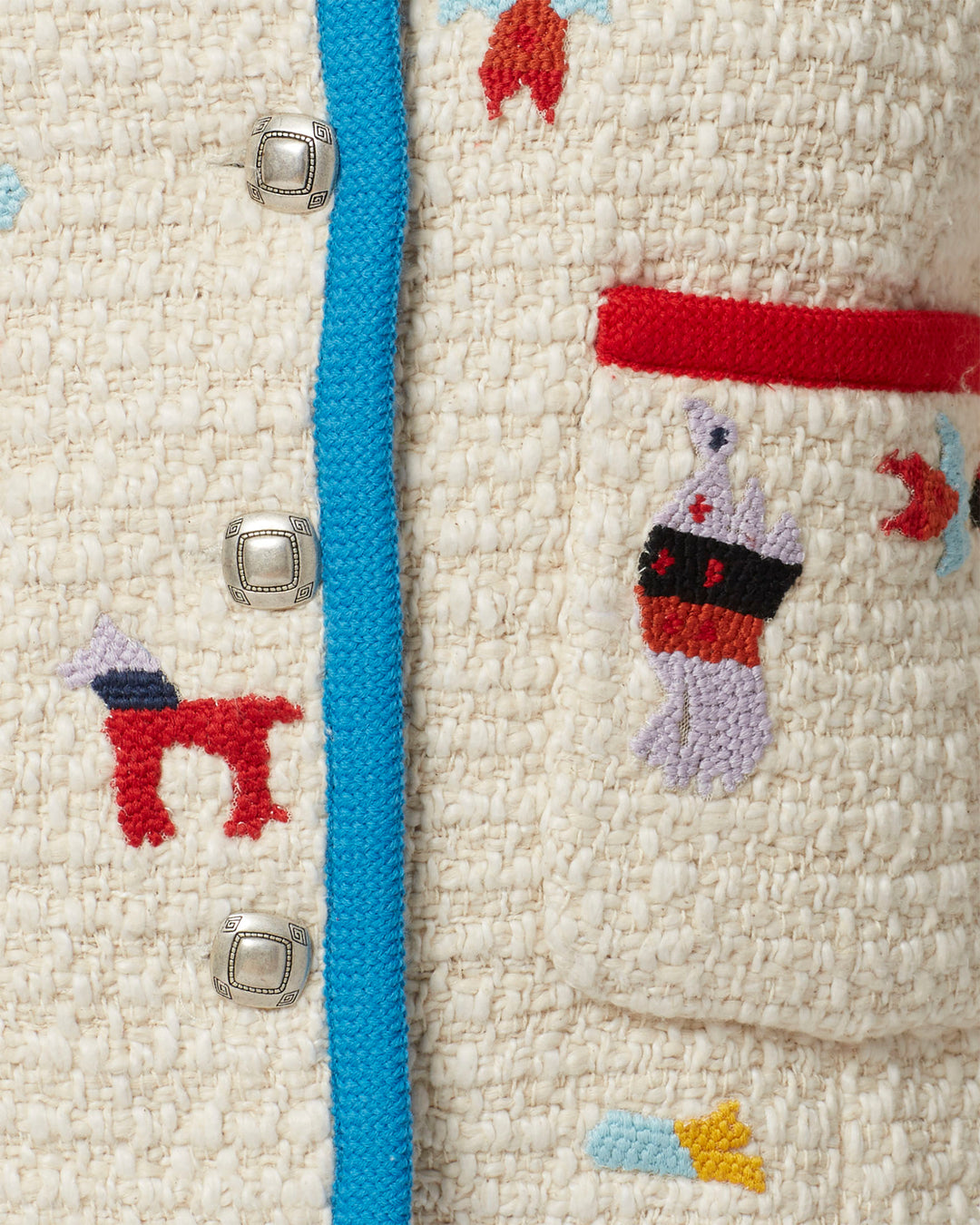 A detailed look at the Rowan Pearl Boucle Jacket reveals vibrant embroidered designs with small animal-like figures. This textured piece is highlighted by a bright blue trim and silver buttons, with a visible pocket showcasing red and black accents.