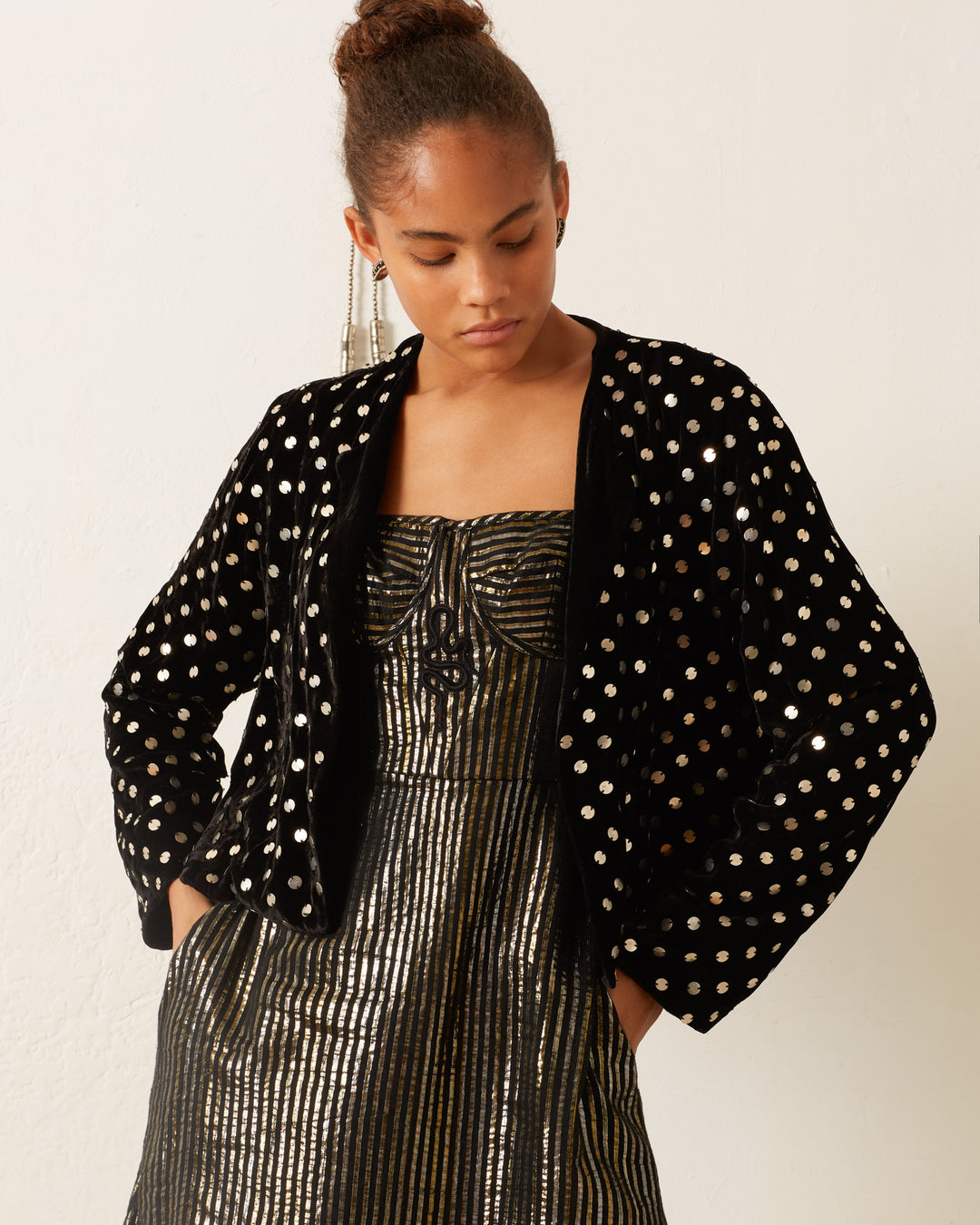 A person stands gracefully against a white background, clad in the Rowan Stardust Velvet Jacket adorned with silver polka dots over a metallic striped dress.