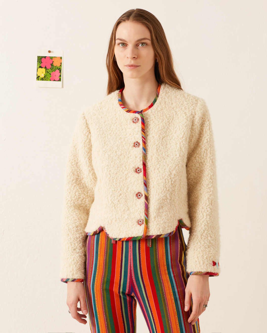 A person stands indoors wearing the Rowan Suri Wool Jacket in cream, which is hand-finished and adorned with colorful buttons, along with multicolored, vertically striped pants. The jacket features a touch of Guatemalan ikat trim, while a small, vibrant artwork hangs on the wall in the background.