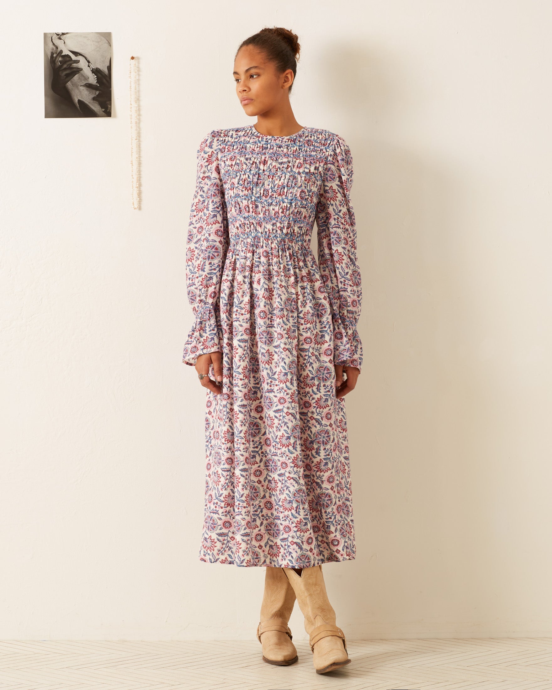 A person stands in the Sabine Amazon Floral Dress, a vintage-inspired piece with long sleeves, an ankle-length cut, a hand-smocked bodice, and a flared skirt. They complement the look with tan boots and have their hair styled in a bun. A black-and-white photo and a string of beads decorate the wall behind them.