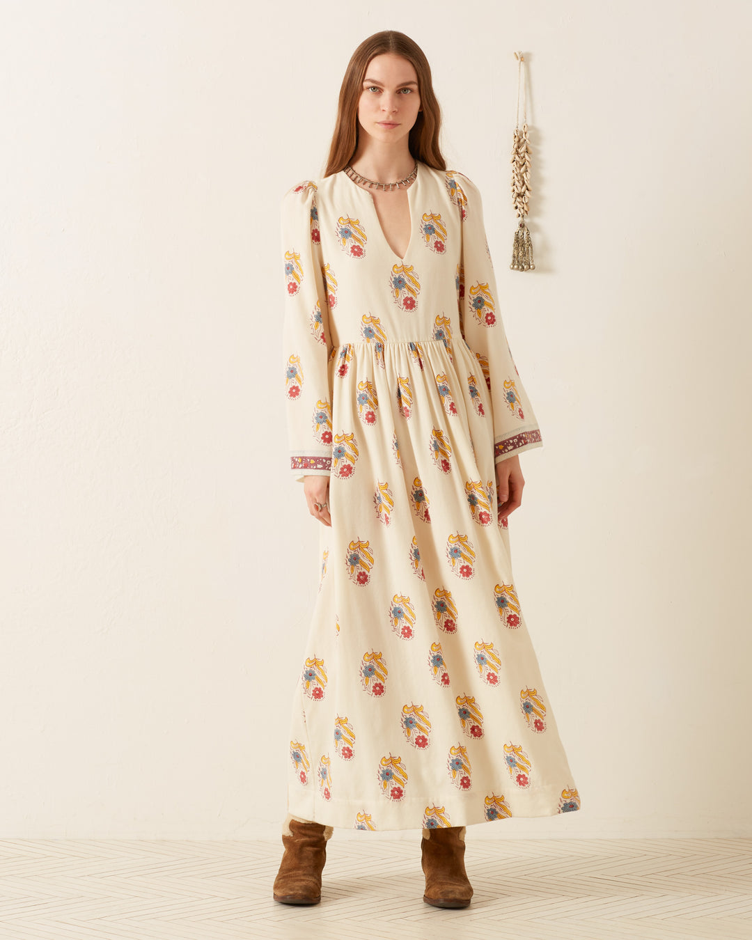 The person is wearing the Savannah Fireflower Wool Dress, featuring a vibrant floral motif with graceful bell sleeves, standing on a light-colored floor. Complemented by brown boots and a beaded necklace, they pose elegantly as a small decorative wall hanging adorns the light wall behind.