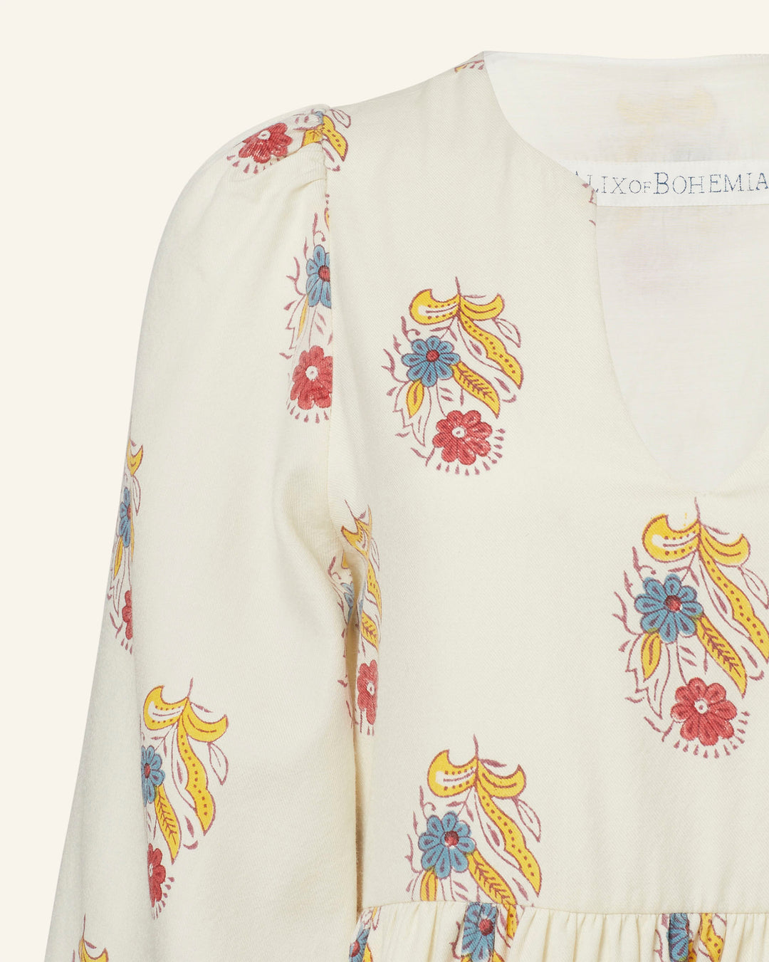 A close-up of the Savannah Fireflower Wool Dress showcases a cream-colored fabric adorned with a vibrant bohemian floral motif in red, blue, and yellow. The dress features a V-neckline and has subtle gathering at the waist for a flowy look, beautifully highlighting the charm of hand-block printing.