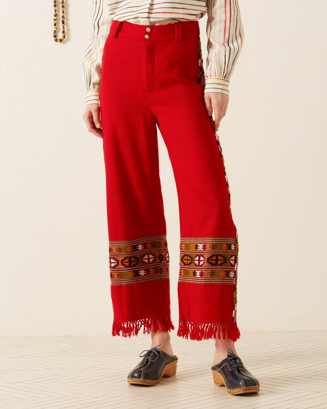 A person wearing the Scout Scarlet Blanket Pant, featuring a bright red color and fringed hem inspired by the Himalayan weaving tradition, pairs their outfit with a striped blouse and wooden platform shoes.