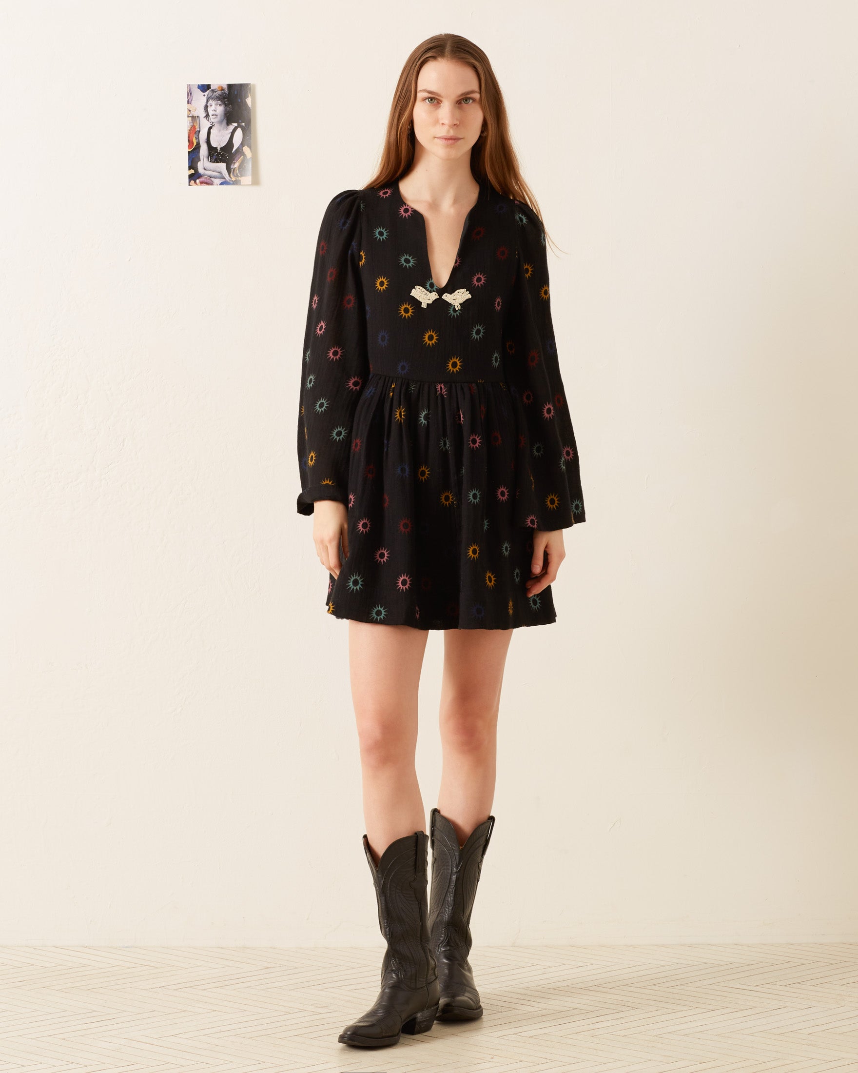 A person with long hair wearing a Silvie Evening Star Dress, hand-embellished with colorful patterns, and black cowboy boots stands in a room. A small poster is on the wall behind them.