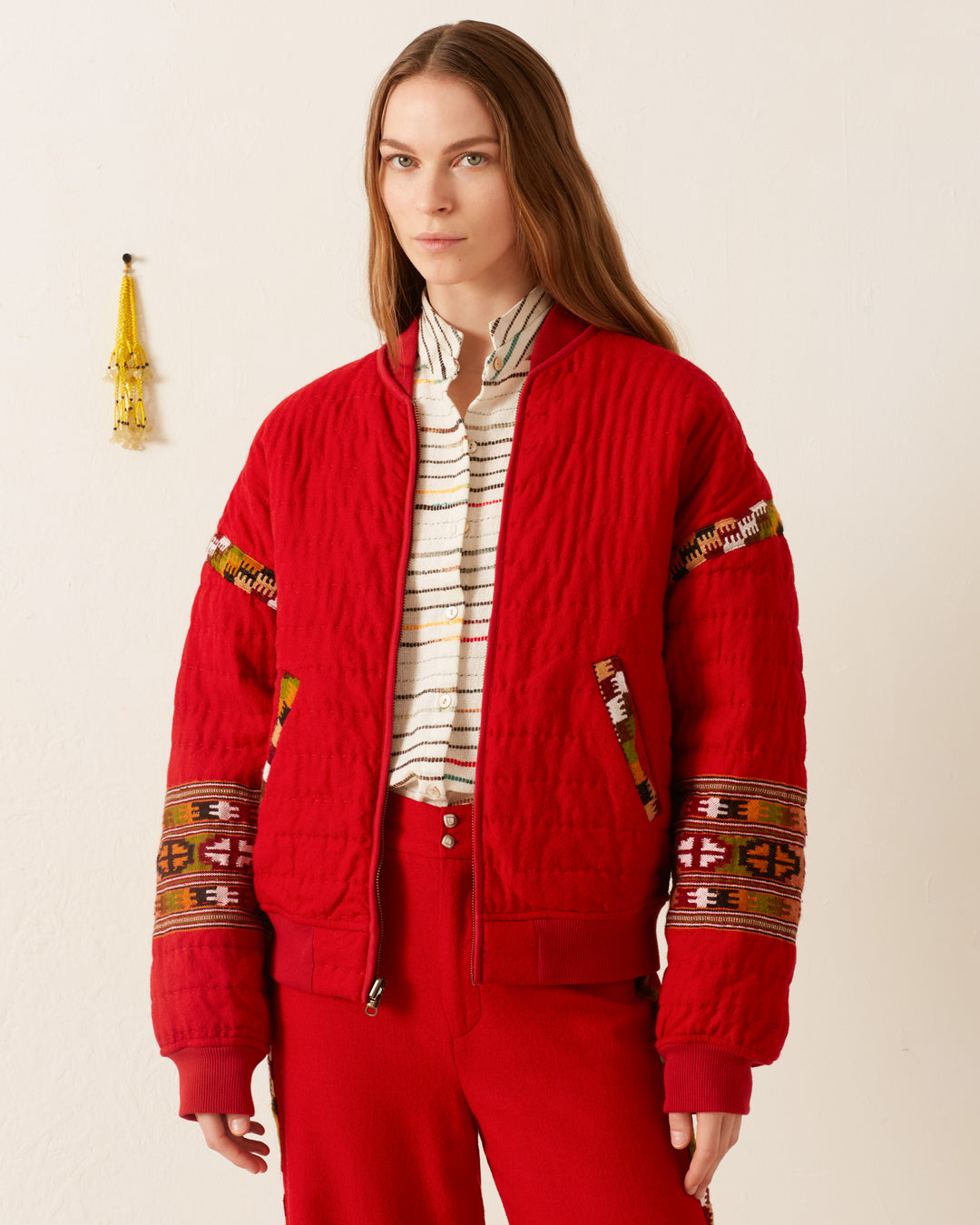 A person stands against a plain wall wearing the Sutton Scarlet Blanket Bomber, featuring intricate hand-quilted patterns, paired with red pants. A small tassel decoration hangs on the wall, enhancing the cozy atmosphere reminiscent of a handwoven Kullu blanket.