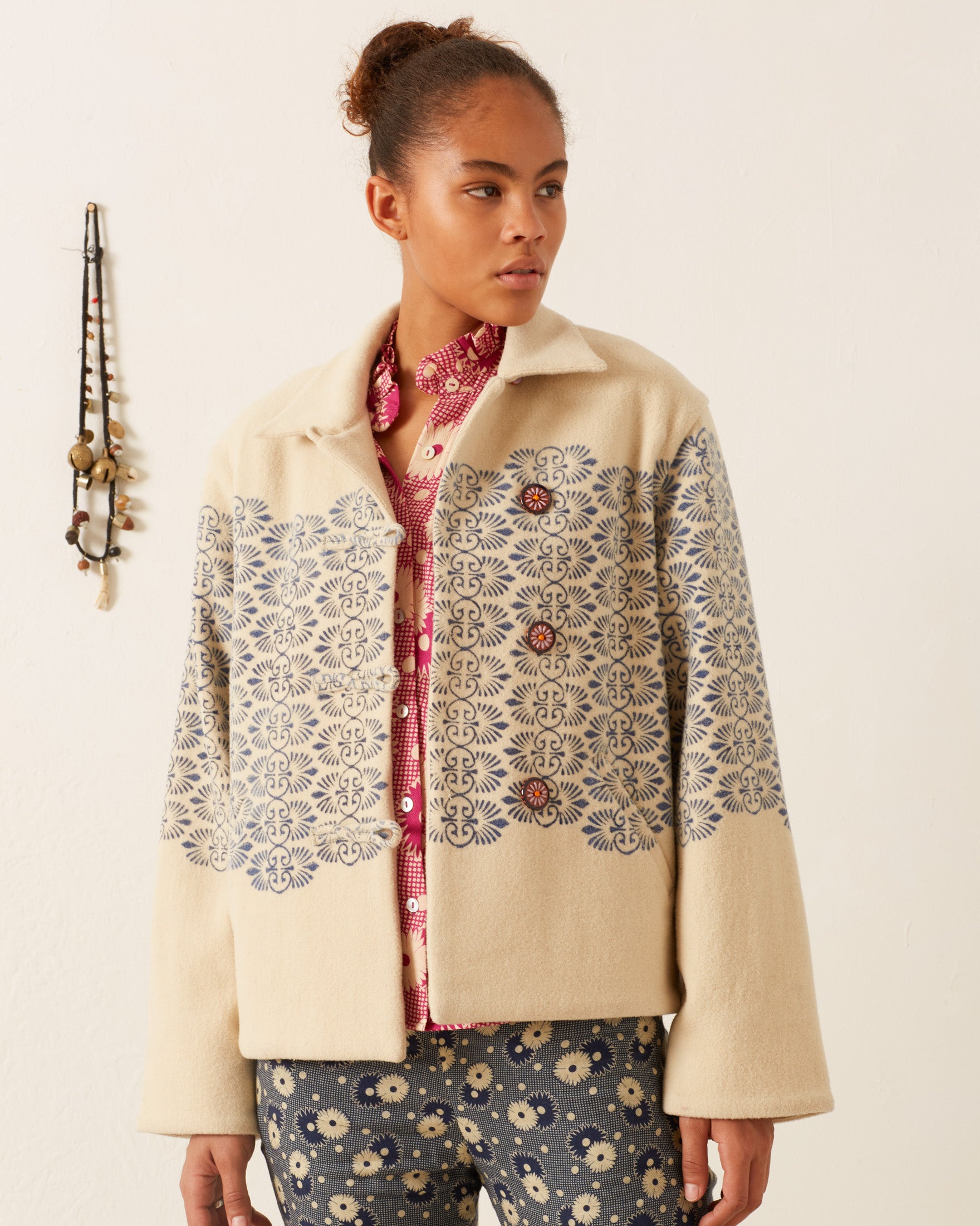 A person with short, curly hair stands indoors wearing a Western Lapis Trellis Jacket over a red patterned shirt. Their hand-block printed blue pants feature a beige floral design. In the background, a decorative item hangs on the wall, adding charm to the scene.