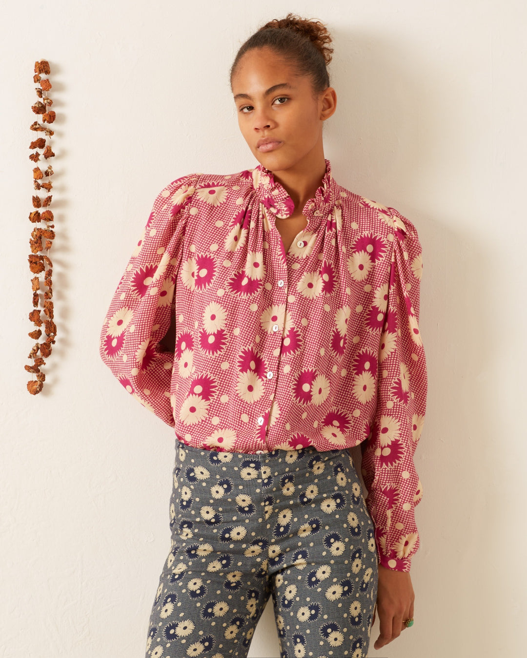 A person stands against a cream-colored wall wearing the Annabel Berry Daisy Shirt and blue floral pants, looking calmly at the camera. To the left, a vertical string of small brown objects is hanging on the wall.