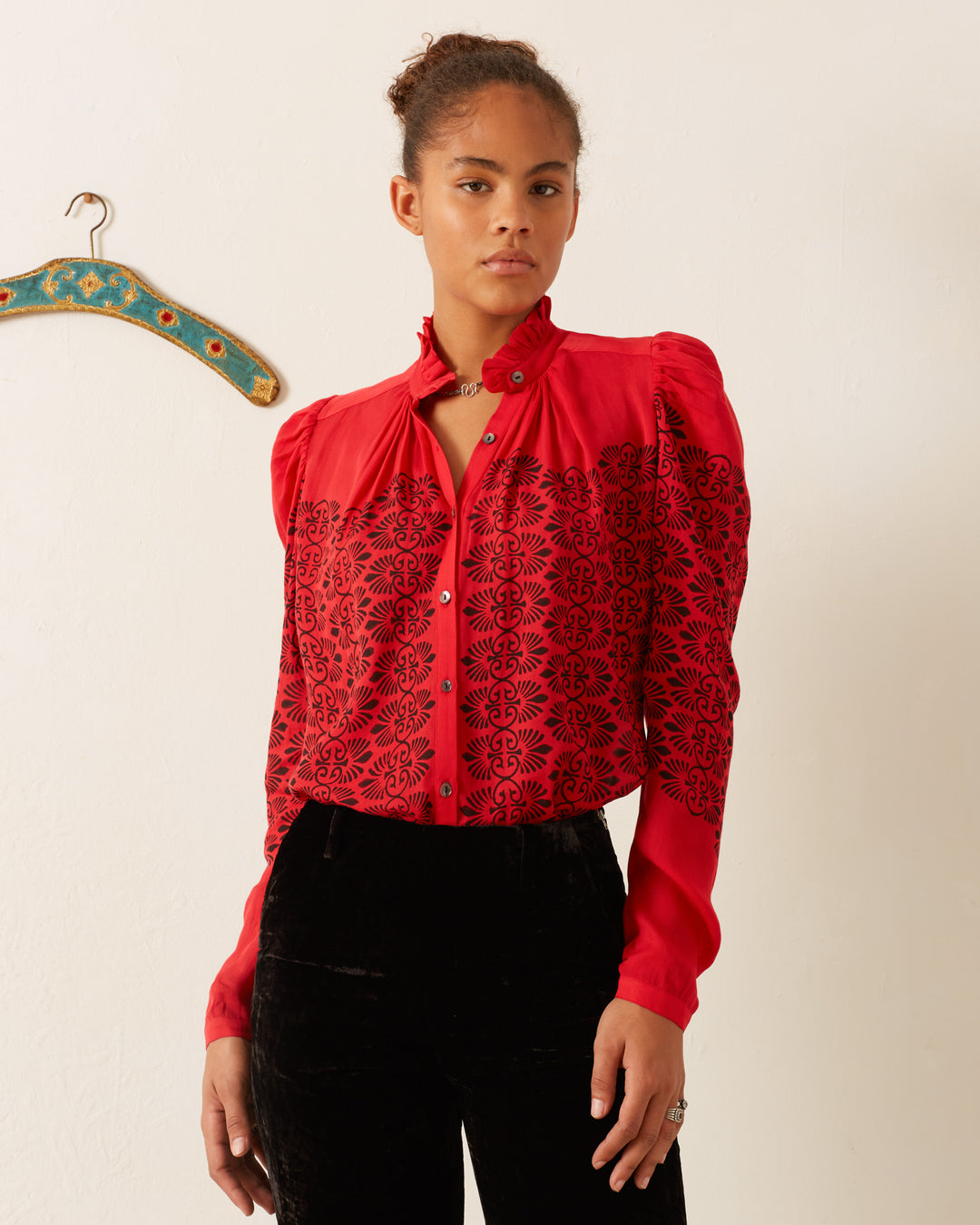 A person wearing the Artemis Cherry Trellis Shirt, designed with vintage-inspired black patterns on a red background, stands against a plain wall. Next to them, a decorative hanger is mounted on the wall. The person is looking toward the camera.