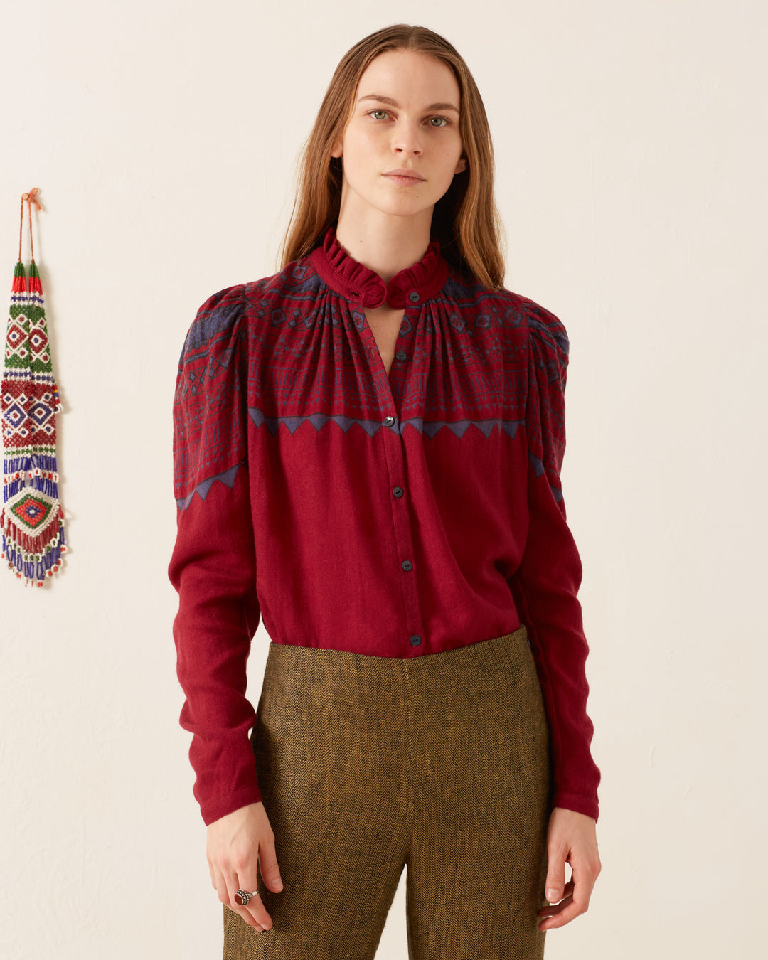 A person with long hair dons an Artemis Plum Fairisle Shirt, a vintage-inspired maroon blouse featuring blue embroidery, paired with brown pants, standing against a plain background. A colorful woven decoration adorns the wall.