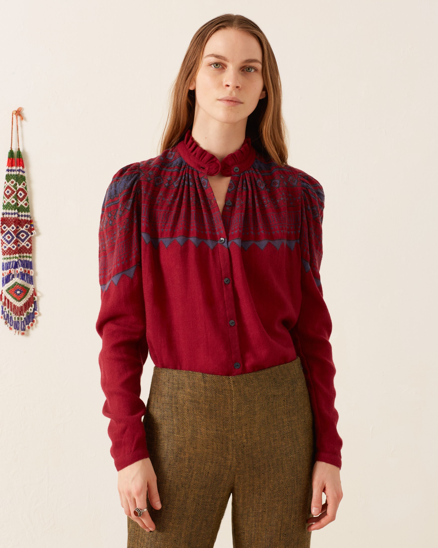 A person with long hair dons an Artemis Plum Fairisle Shirt, a vintage-inspired maroon blouse featuring blue embroidery, paired with brown pants, standing against a plain background. A colorful woven decoration adorns the wall.