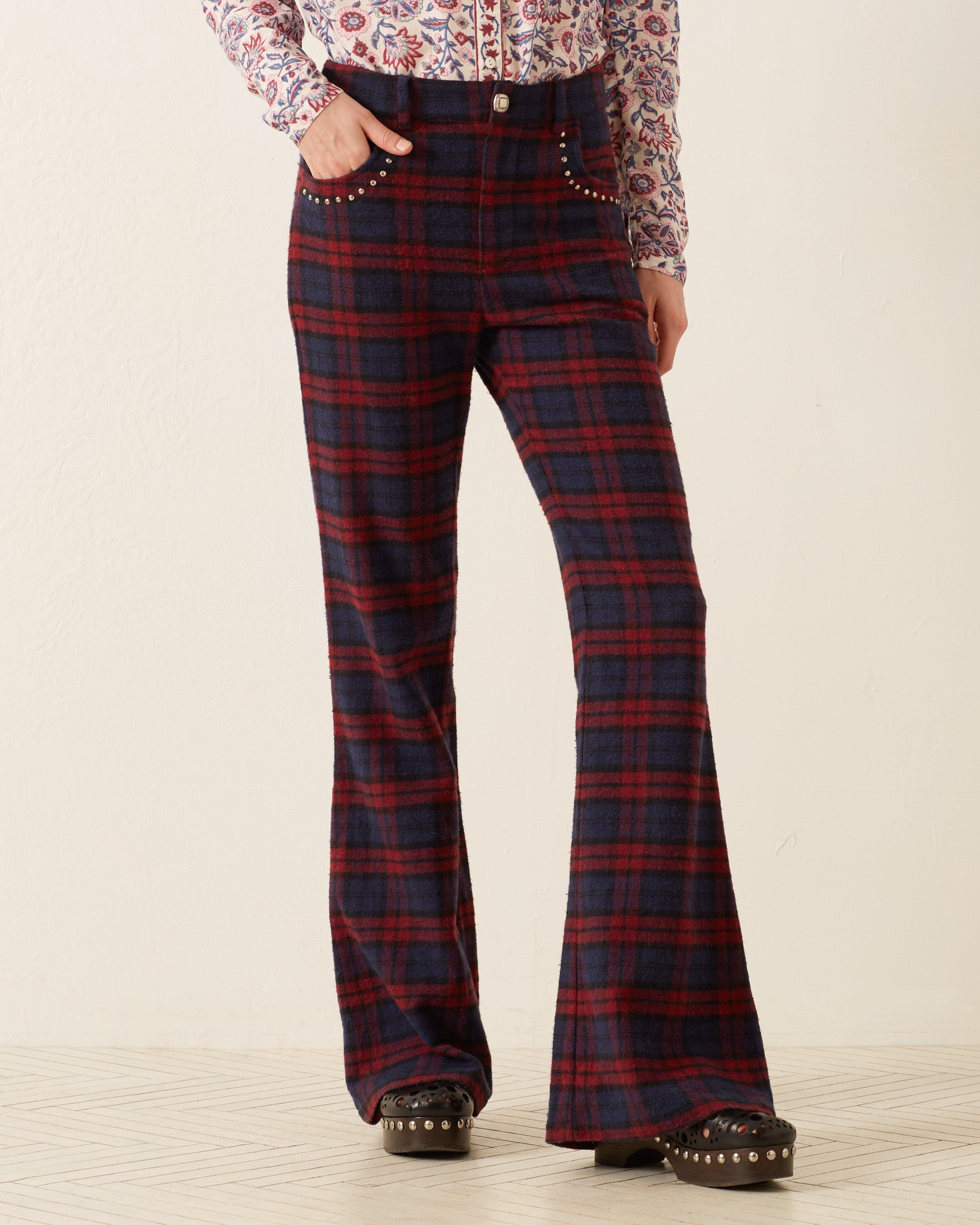 A person is wearing high-waisted, Bunny Berry Plaid Pants in red and black tartan, featuring studded pockets, paired with a floral blouse. Their stylish look is completed with hand-embellished details as they stand confidently on a light-colored floor, showcasing studded black shoes.