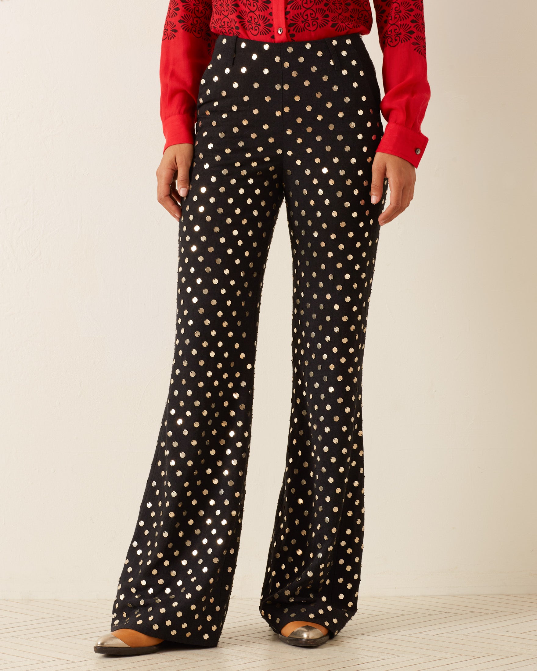 A person wearing the Charlie Stardust Pants in black, adorned with a gold polka dot pattern and silver sequins, paired with a red long-sleeved shirt. These high-waisted pants feature flared legs. The outfit is completed with shoes that have pointed toes and metallic tips, set against a neutral background with light-colored flooring.