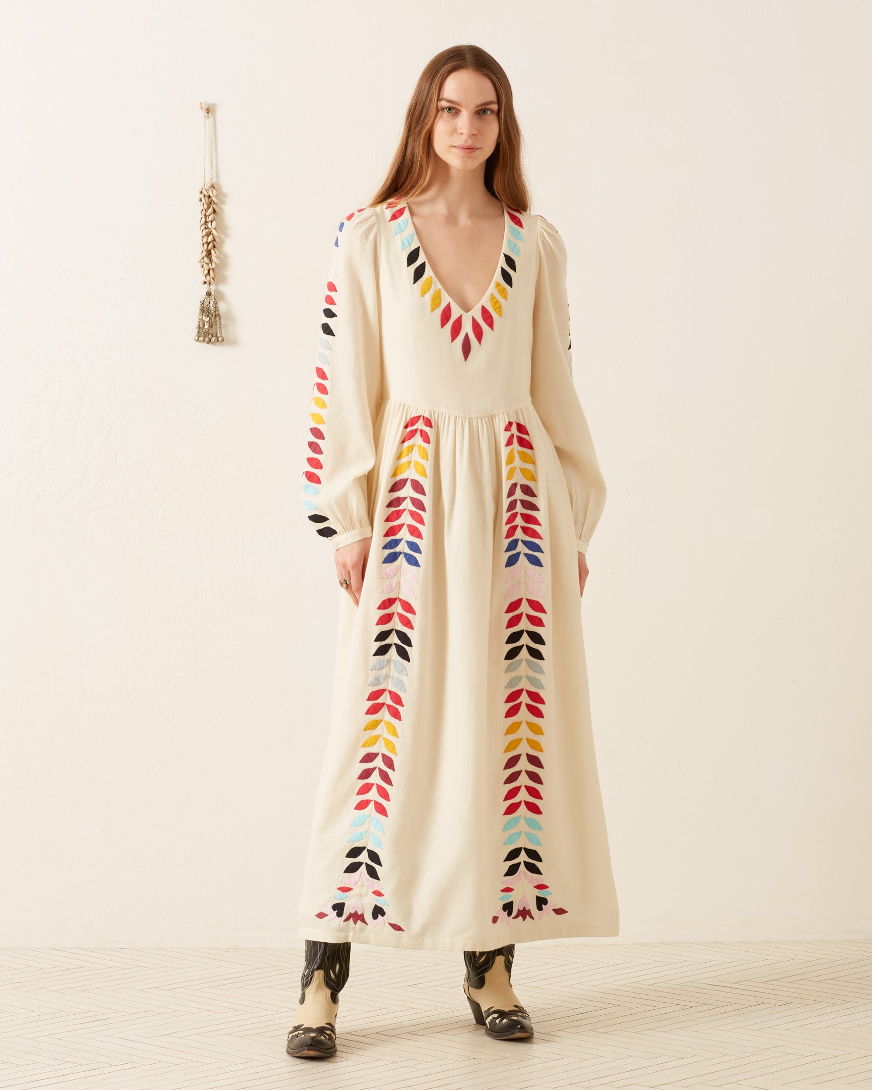 A person stands in the Imogen Ivory Prism Dress, showcasing colorful geometric patterns with a V-neck and long sleeves. The ensemble is complemented by black boots and set against a plain, light-colored wall with a decorative tassel hanging.