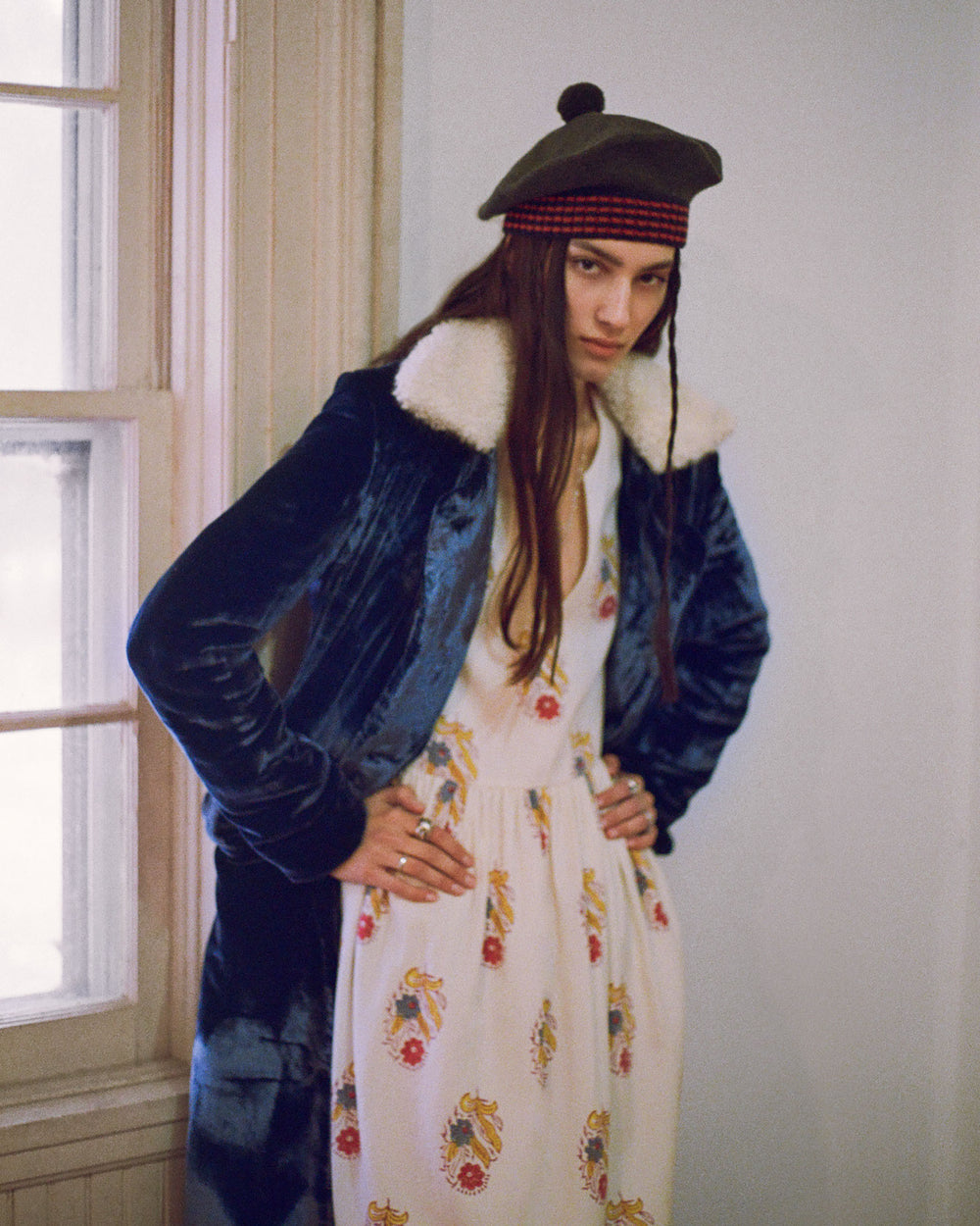 A person wearing a dark coat with a white fur collar, the Savannah Fireflower Wool Dress featuring a hand-block printed floral motif, and an olive green beret with a red band stands with hands on their hips next to a window. The room's neutral-colored walls create a serene backdrop.