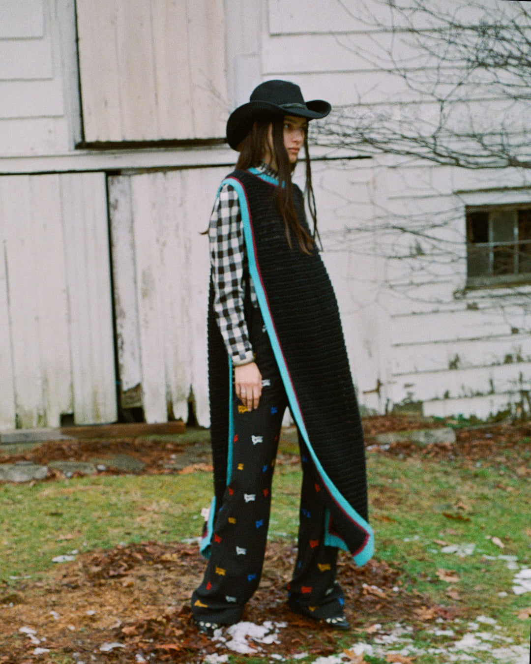 A person wearing a black hat, plaid shirt, Omer Equinox Crochet Top with blue trim, and patterned pants stands outside near a white wooden building.