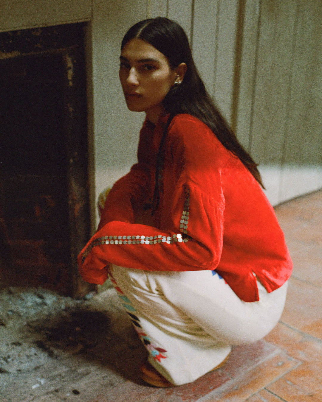 A person is squatting near a fireplace, dressed in the Dylan Ruby Velvet Tunic with patterned details and white pants adorned with designs.