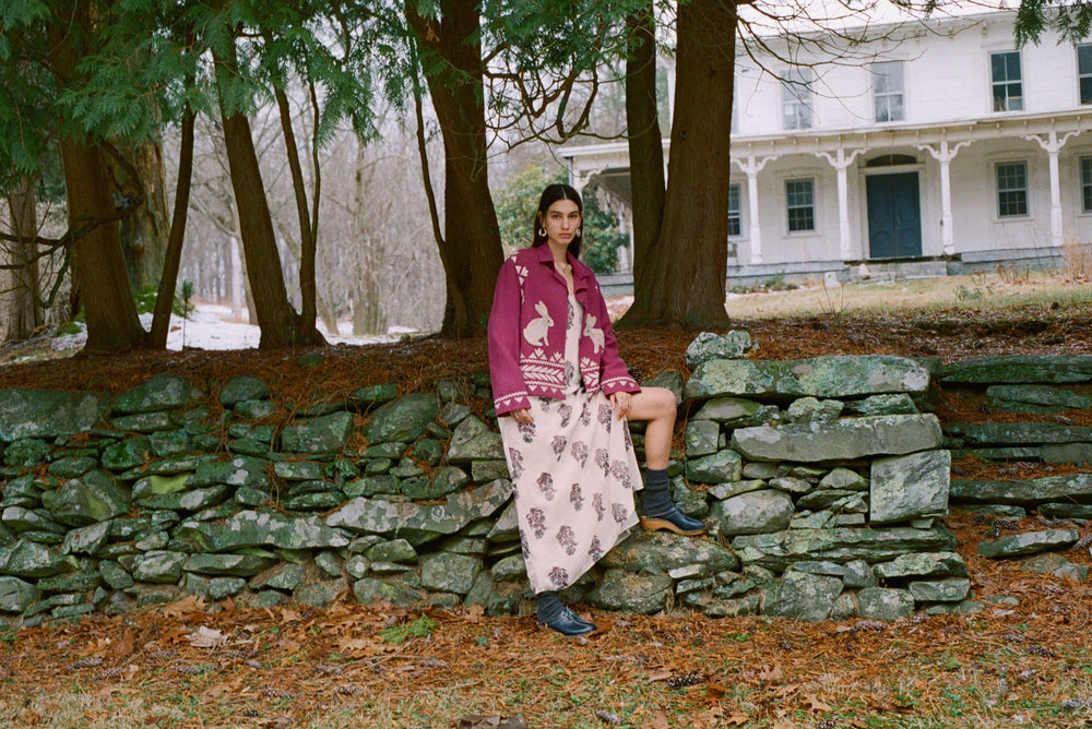 A person sits on a stone wall in a wooded area wearing the Paloma Azure Mirabilis Dress along with a maroon sweater that features animal patterns and black boots. A large house can be seen in the background.