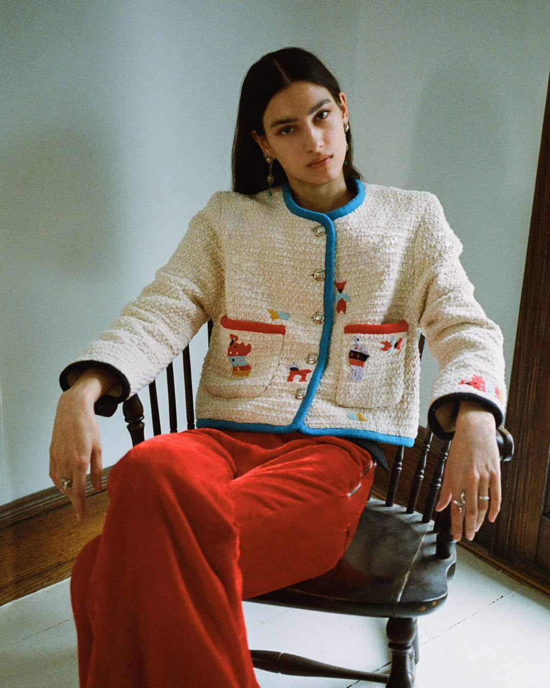 A person with long dark hair sits in a wooden chair, wearing the Rowan Pearl Boucle Jacket featuring colorful pocket details and red pants. The room is decorated in light, neutral tones, and the soft natural lighting enhances their relaxed pose.