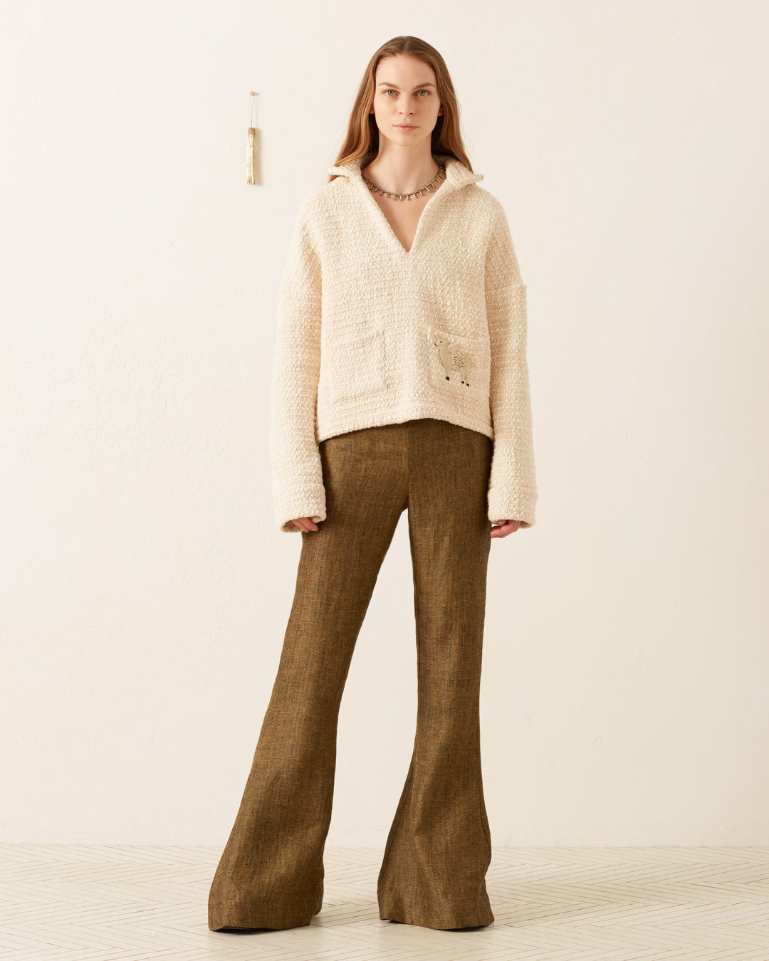 A person stands against a plain background, wearing the Allegra Pearl Bouclé Top featuring handmade llama appliqué and brown flared pants.