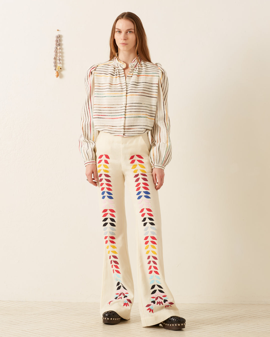 A person wearing a striped blouse and the Charlie Ivory Prism Pant, featuring '70s-style wool with colorful patterns and floral appliqué, stands against a plain wall.