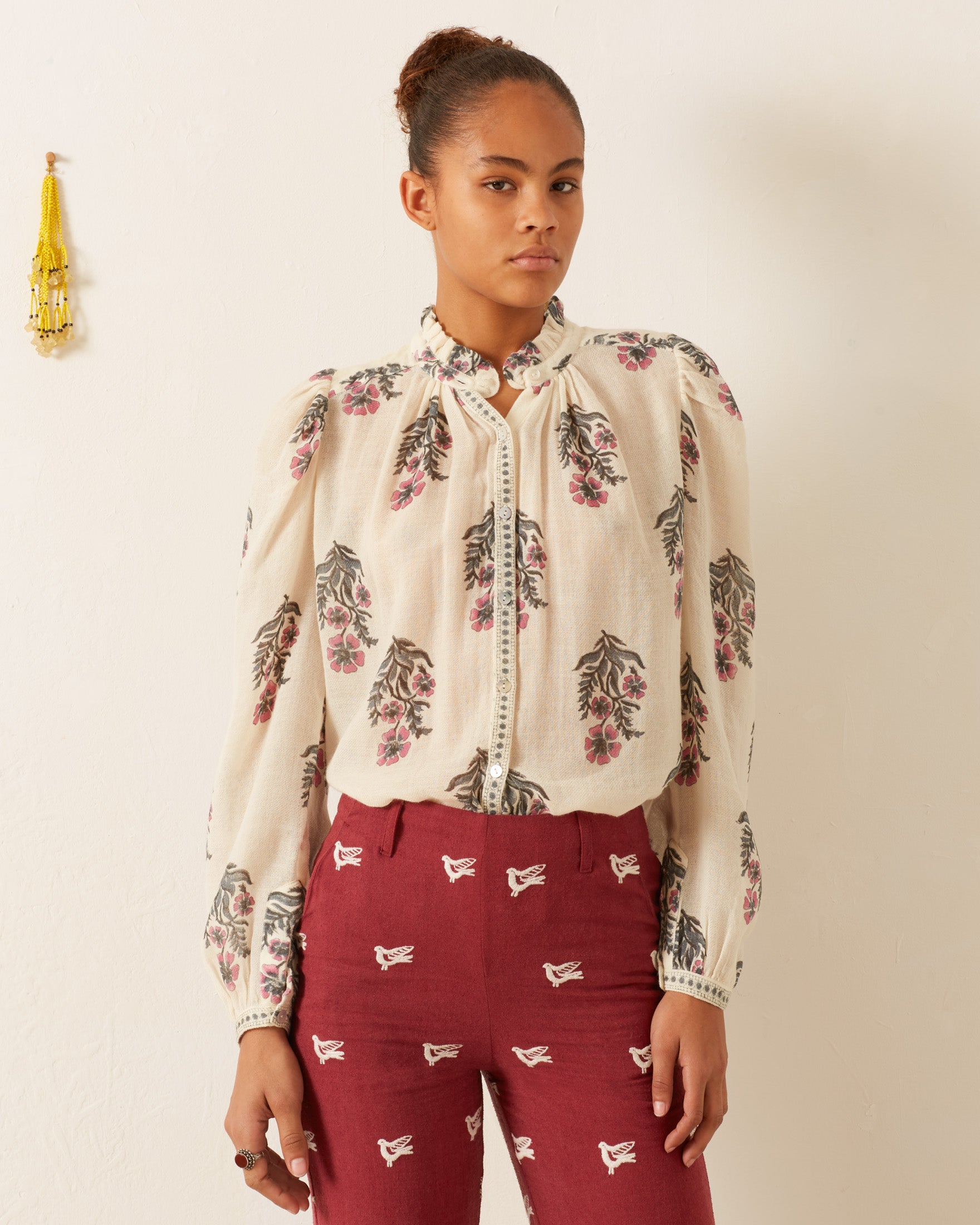 A person stands against a white wall, wearing the Annabel Mirabilis Wool Shirt with a floral pattern and maroon pants featuring bird embroidery. A tassel decoration adorns the wall. Their hairstyle is an updo, and they wear a neutral expression.