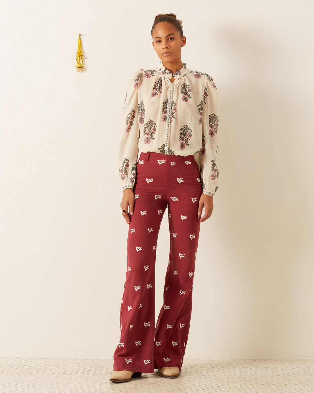 A person stands against a white wall wearing the Annabel Mirabilis Wool Shirt with a vintage-inspired floral motif, paired with red patterned pants. The floor is light wood, and a decorative yellow item on the wall in the background adds to the charming scene.