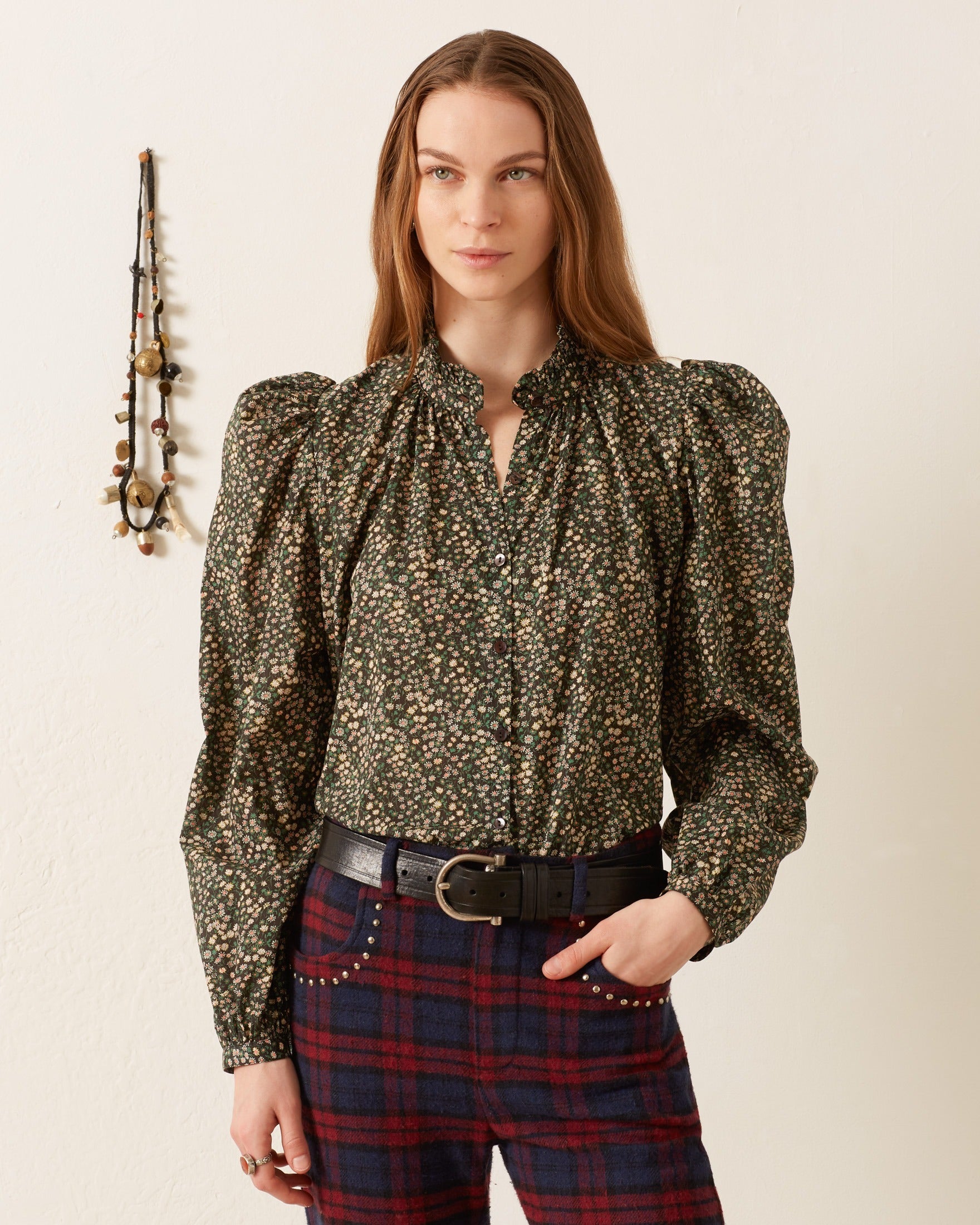 A person wearing the Annabel Moonflower Shirt, a vintage-inspired green ditsy floral blouse with puffed sleeves, paired with red plaid pants stands against an off-white wall adorned with a decorative hanging featuring beads and tassels.