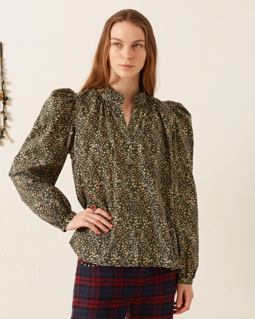 A person with long hair is wearing the Annabel Moonflower Shirt, a vintage-inspired blouse with ditsy floral patterns and puffed sleeves, along with plaid pants. They stand against a plain background, looking directly at the camera with one hand on their hip.