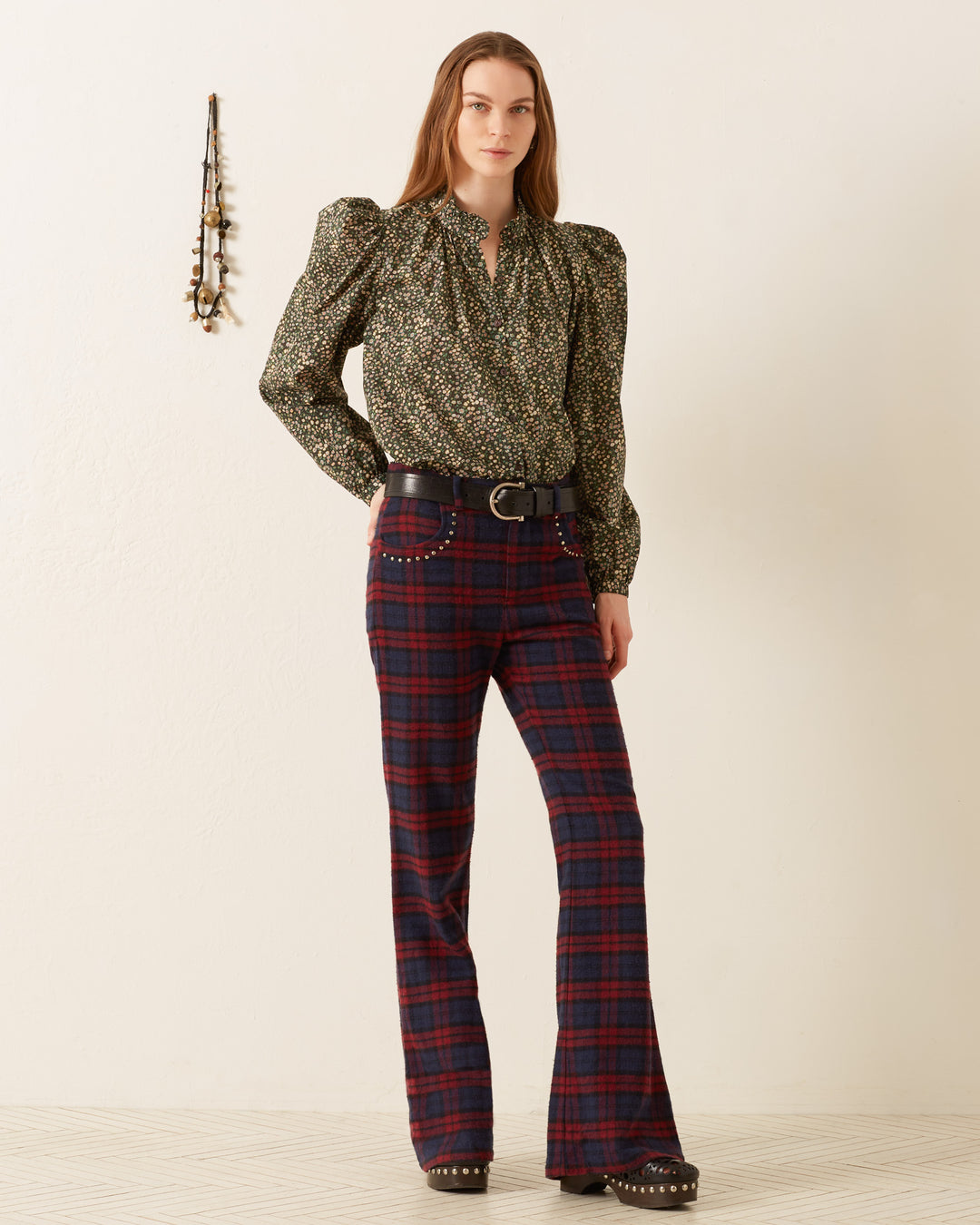 A person wearing the Annabel Moonflower Shirt, featuring a ditsy floral print and long sleeves, pairs it with red plaid pants against a plain wall. They are also sporting black studded shoes and a black belt. In the background, a necklace hangs on the wall, enhancing the vintage-inspired ambiance.