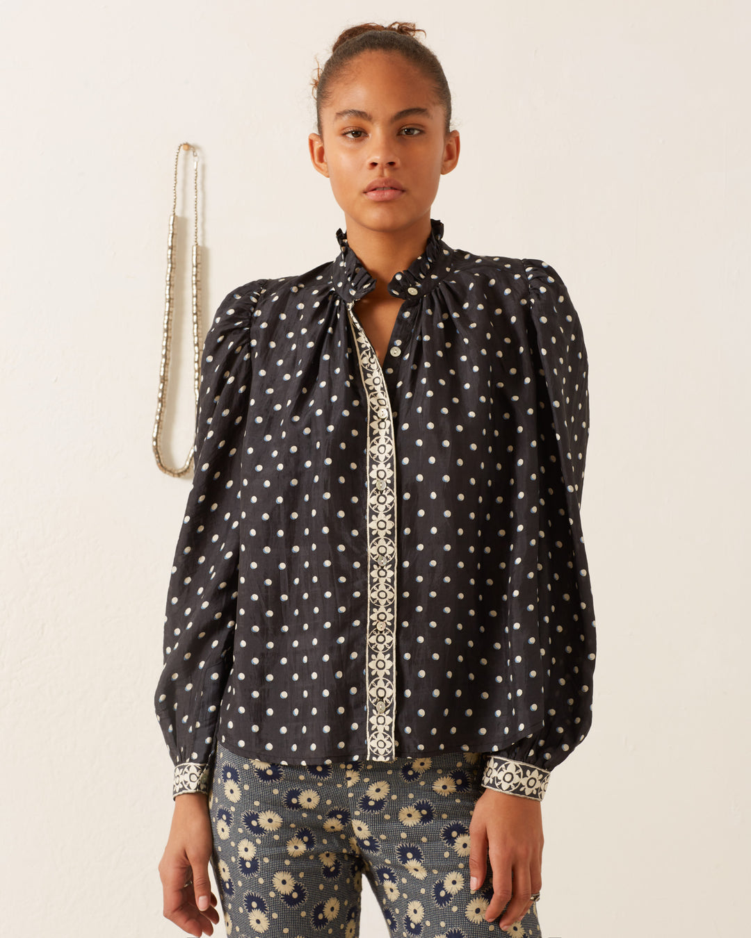 A person stands against a plain background, wearing the Annabel Raven Dot Silk Shirt. This blouse features intricately patterned cuffs, slightly puffed long sleeves, and hand-block printed decorative trim around the neckline and down the front.