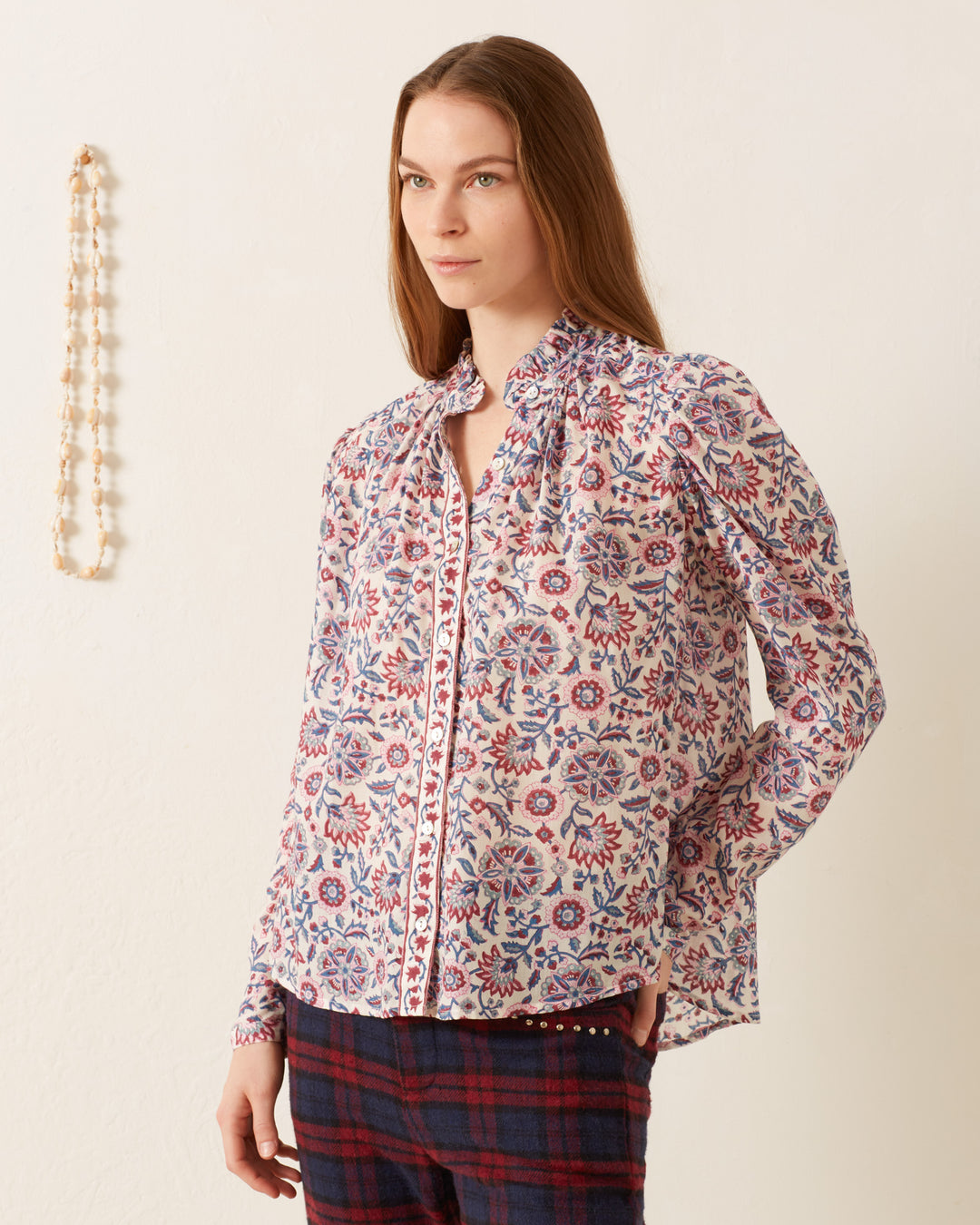 A woman stands against a cream-colored wall, wearing the Artemis Amazon Floral Shirt with intricate vintage-inspired designs and plaid pants. A beaded necklace hangs on the wall beside her.