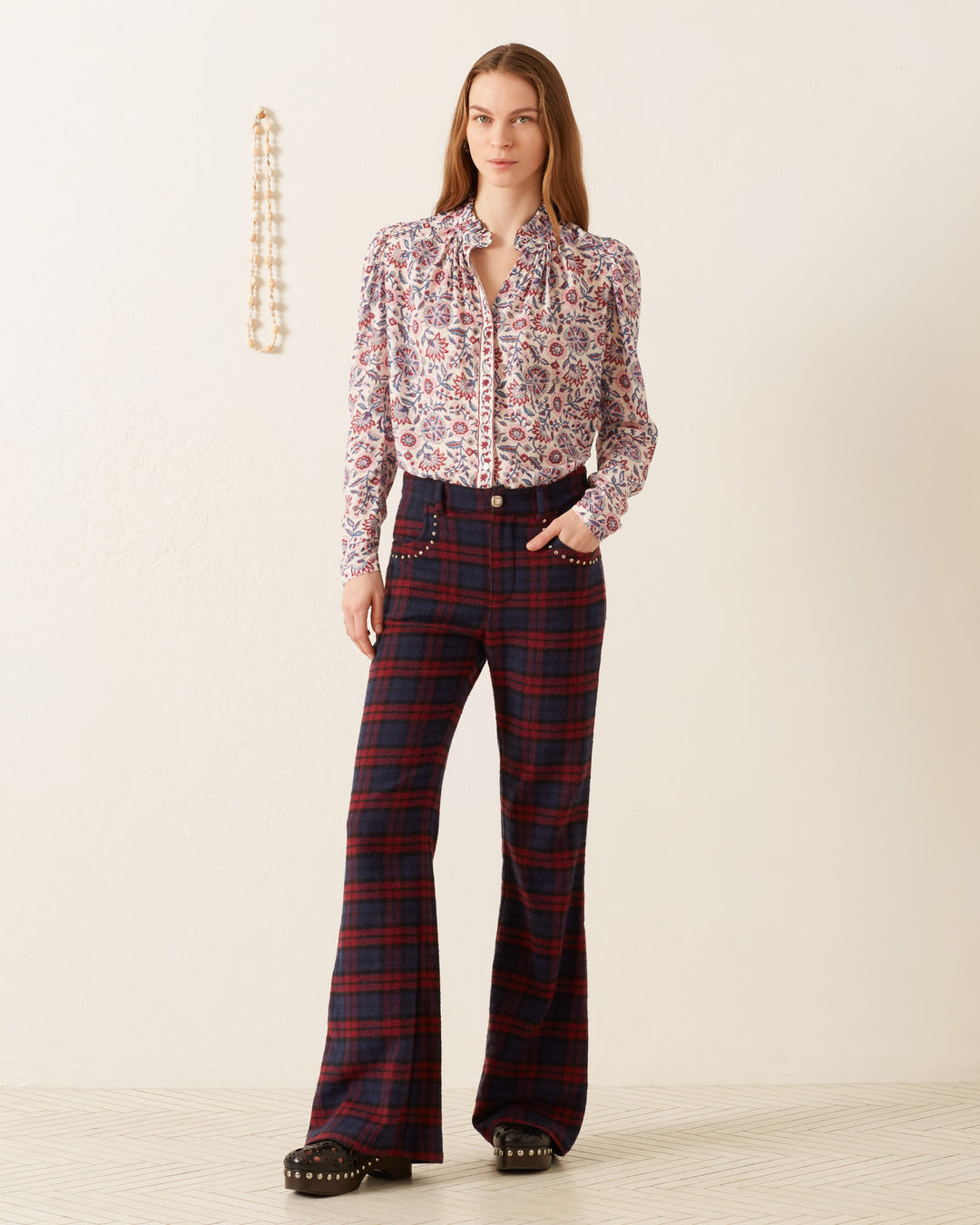 A person stands against a plain wall, wearing an Artemis Amazon Floral Shirt paired with plaid trousers. They hold one hand in their pocket and wear platform shoes. A string of beads hangs on the wall behind them.