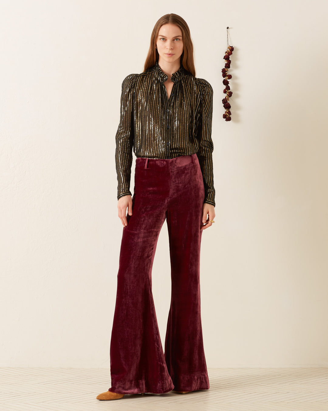 A woman stands in a minimalist room, exuding '70s disco flair with her shimmering Artemis Moonlight Shirt featuring metallic stripes and burgundy velvet bell-bottom pants. Brown shoes peek out underneath, while a dried flower arrangement whimsically adorns the wall beside her.