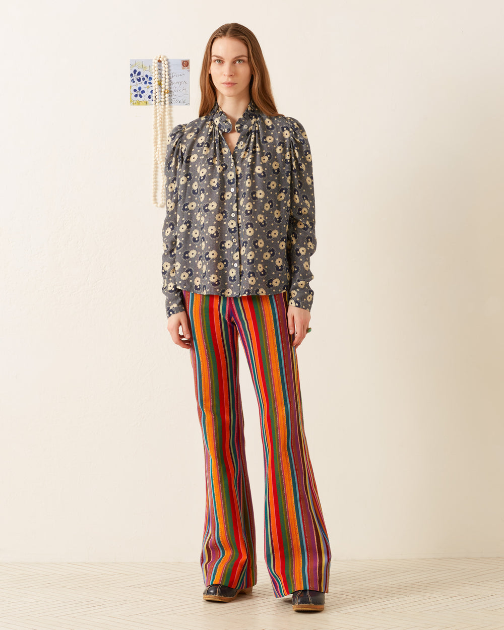 A person stands in a room wearing the Artemis Navy Daisy Shirt, featuring a vintage-inspired ’70s-style floral print, paired with colorful striped flared pants. A necklace and a small piece of art adorn the wall behind them, while the floor showcases a light pattern that completes the retro aesthetic.