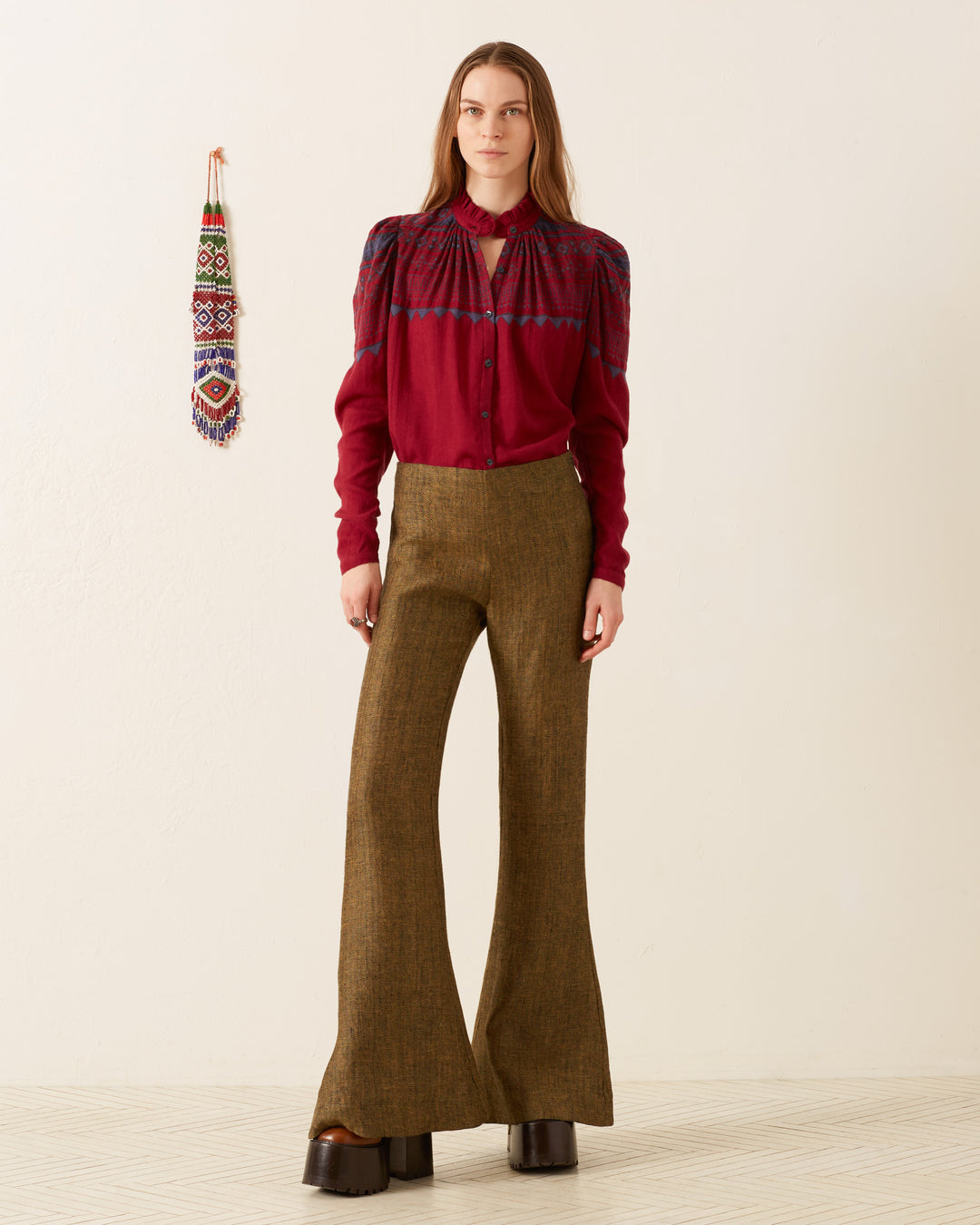 A person stands against a plain background wearing the Artemis Plum Fairisle Shirt, a vintage-inspired design with intricate patterns, paired with brown flared trousers. Beside them, the wall showcases a hand-block printed, colorful hanging textile ornament.