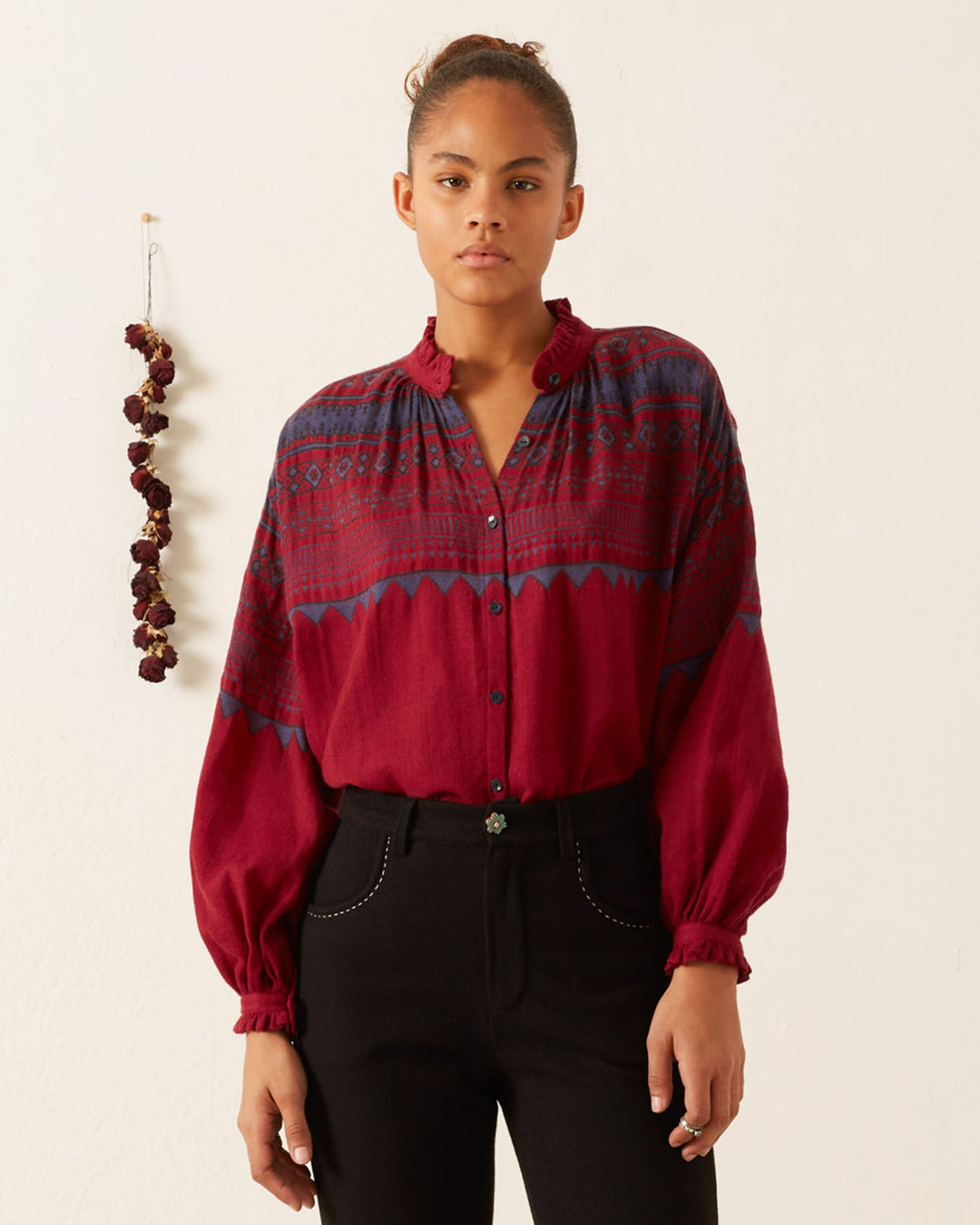 A person standing indoors wearing a Poet Plum Fairisle Blouse paired with black pants, with a garlic braid hanging on the wall in the background.