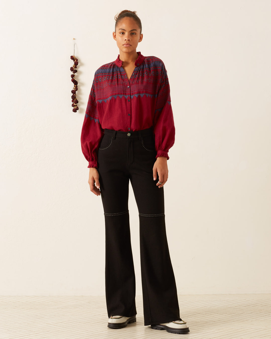 A person stands against a white wall wearing the Poet Plum Fairisle Blouse and black pants. A string of dried peppers hangs on the wall to the left, adding a touch of rustic charm.