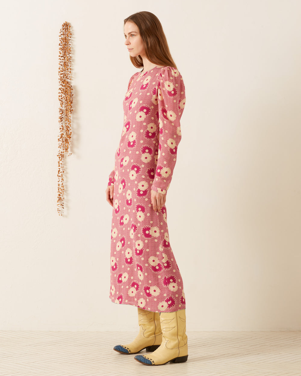 A person with long hair stands in profile, wearing the Astrid Berry Daisy Dress and yellow cowboy boots. They are in a minimalist room with a textured wall and a beaded decoration hanging down one side.