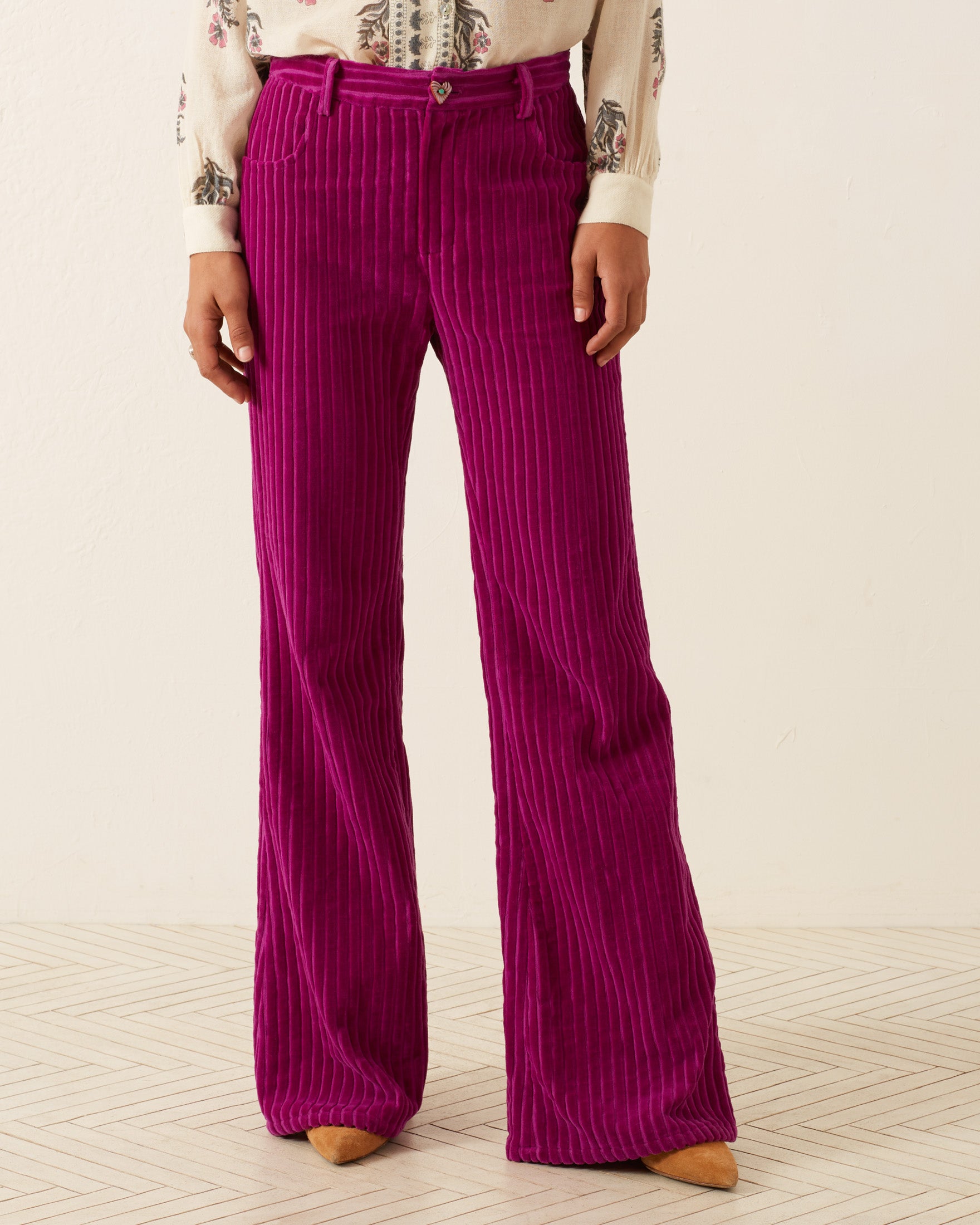 A person stands on a light herringbone floor, wearing vibrant Bunny Violet Velvet Pants paired with a patterned shirt.