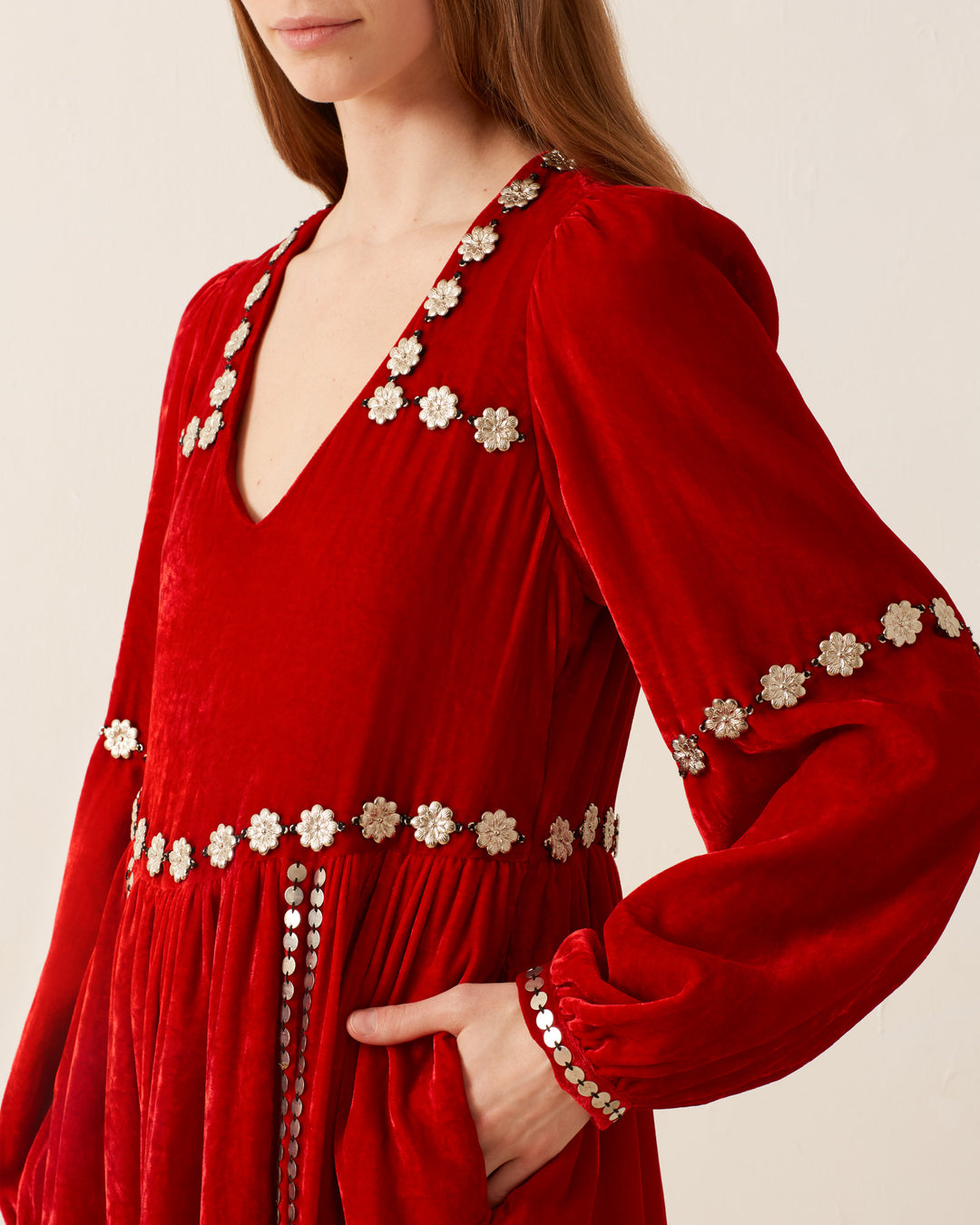 A woman elegantly stands in an Imogen Ruby Velvet Dress, featuring vintage-inspired silver floral embellishments on luxurious red velvet. The gown boasts a V-neckline and a cinched waist, with her hand tucked into a side pocket against the plain, light-colored wall.