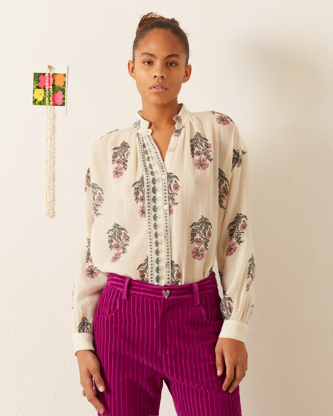 A person stands against a white wall wearing the Kiki Mirabilis Wool Shirt, a vintage-inspired cream blouse with floral patterns, paired with bright pink corduroy pants. A small decorative piece adorned with flowers hangs on the wall beside them, perfectly matching the shirt's elegant floral motif.