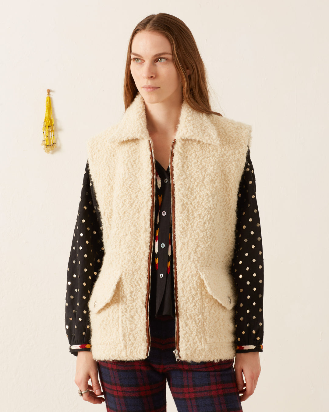 A person with long hair stands against a plain background, wearing a cozy Mickey Suri Wool Vest over a polka dot shirt, with a yellow tassel hanging on the wall.