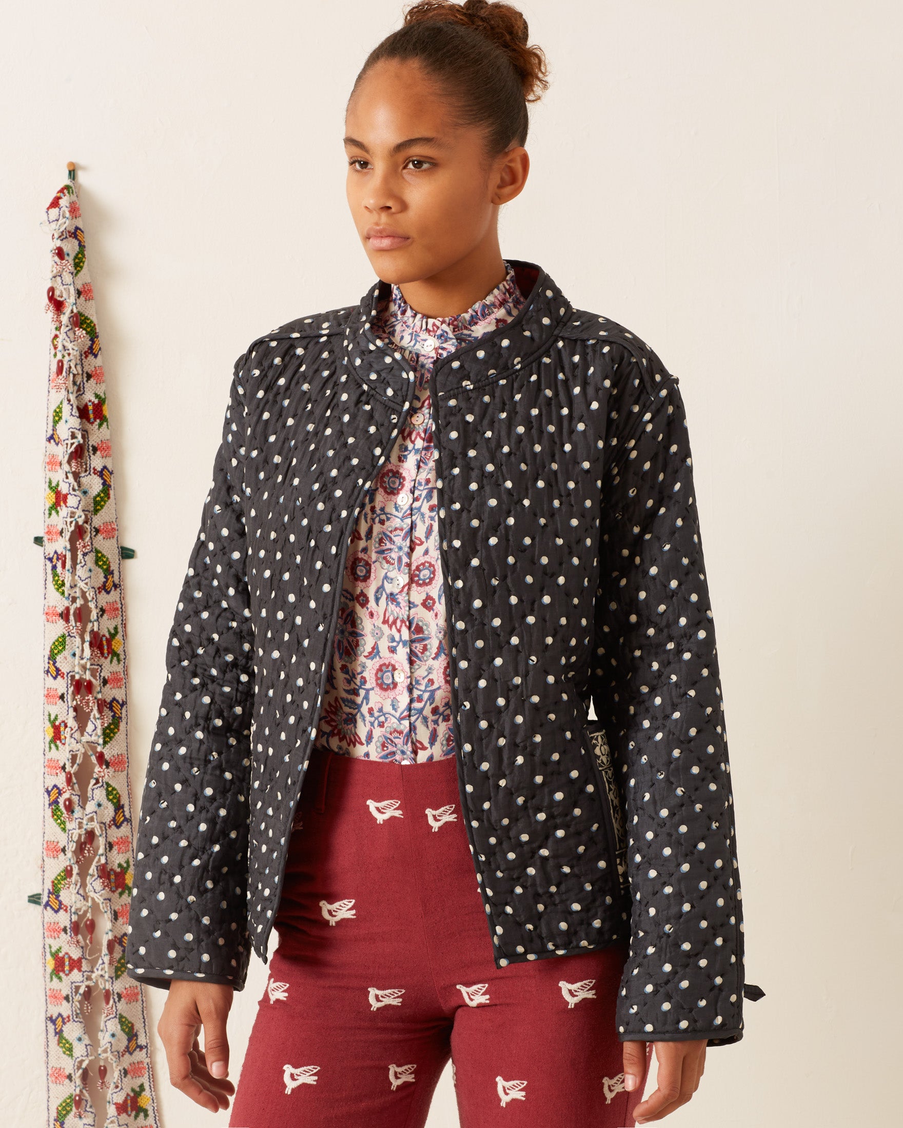 A person wearing a Tamil Raven Dot Silk Jacket, a floral shirt, and red pants stands against a white wall with a floral-patterned fabric hanging beside them.