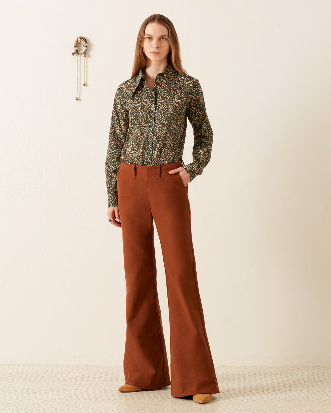 A person stands against a beige wall, wearing the Dolly Moonflower Shirt and brown flared pants, hands in pockets. A small decorative item is on the wall.